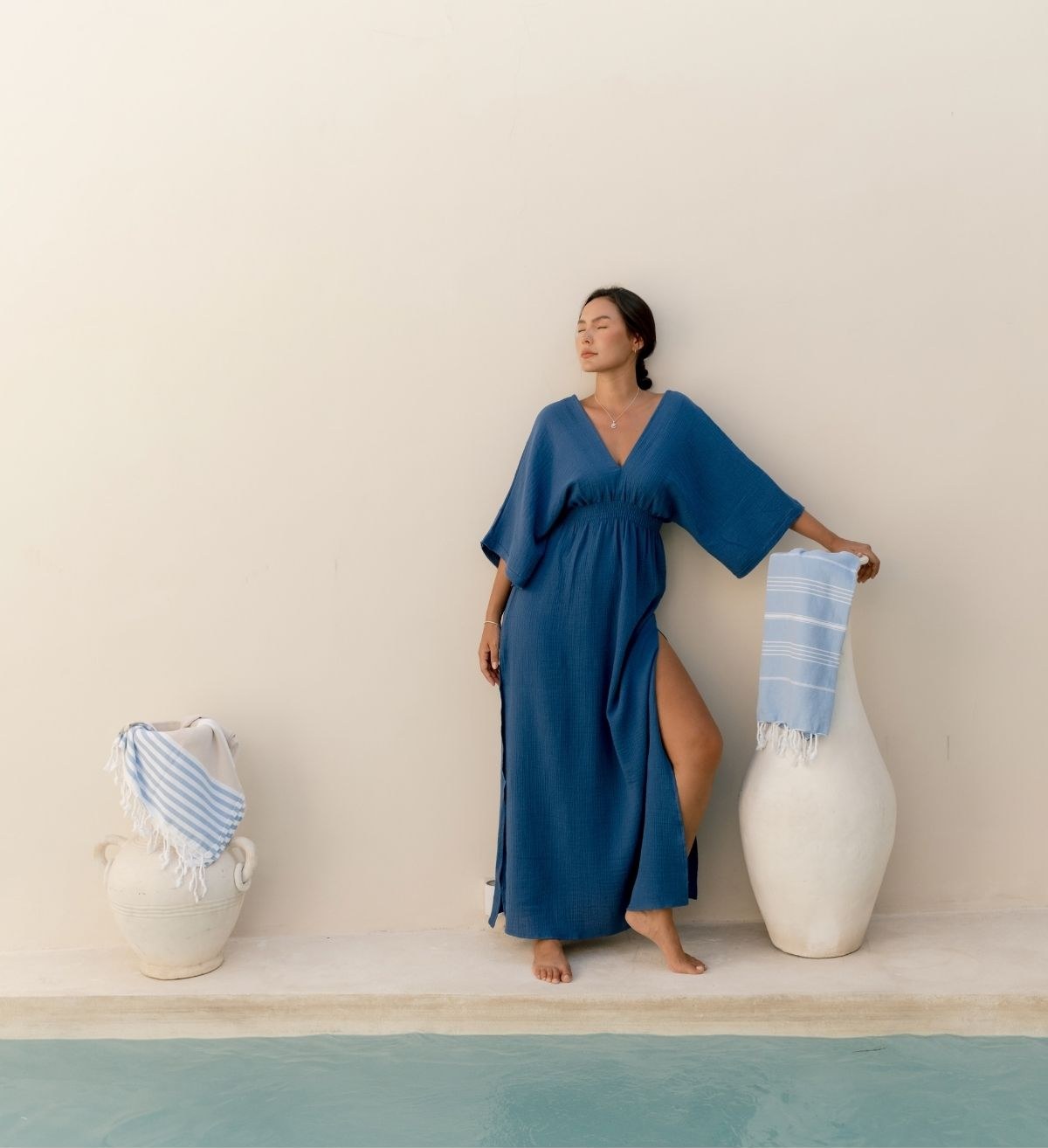 A woman in a blue kaftan beside white vases draped with striped Turkish towels
